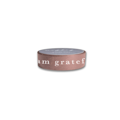 The "I Am Grateful Washi Tape - Mauve" by Maylay Co. is housed in a small, round container featuring a light brown side with the text "I am grateful." Its gray top showcases subtle text and exudes a minimalistic charm, making it perfect for organizing your decorative washi tapes or other scrapbooking supplies. The image is set against a white background for clarity.